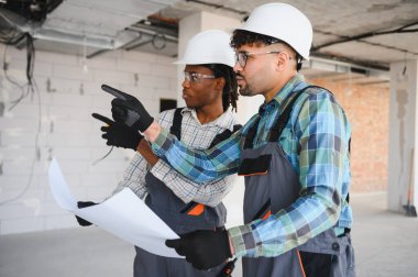 Şapka ve eldiven takmış iki mimar bir planı inceliyor ve bir inşaat alanındaki ilerlemeyi tartışıyorlar.