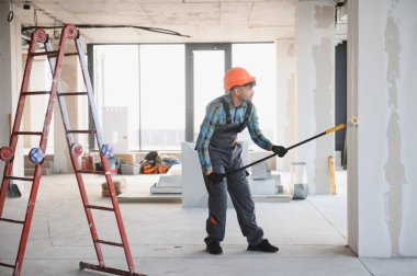 İnşaat işçisi inşaat halindeki bir binanın içinde teleskopik saplı patenli fırçayla duvarları boyuyor.