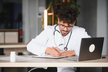 Tıp bürosunda dizüstü bilgisayarla çalışırken not alan odaklanmış erkek doktor, sağlık hizmetleri sunuyor.