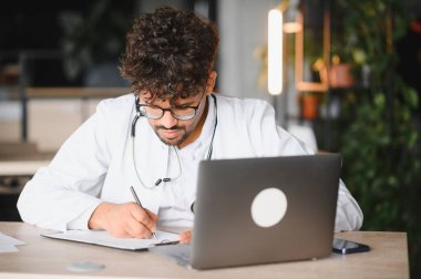 Beyaz önlük ve steteskop giyen odaklanmış erkek doktor modern bir klinikte dizüstü bilgisayar üzerinde çalışırken not alıyor.