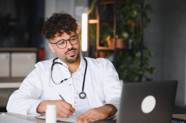 Modern bir ofiste dizüstü bilgisayar üzerinde çalışırken steteskopla tıbbi notlar yazan odaklanmış bir erkek doktor.