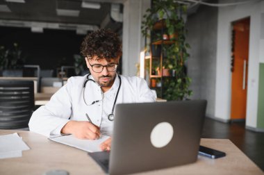 Odaklanmış erkek pratisyen bilgisayar kullanırken not alıyor, modern klinikte çevrimiçi danışmanlık veya tıbbi bilgi araştırması yapıyor