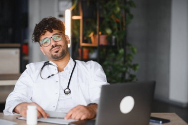 Kendine güvenen Arap erkek doktor masasında oturuyor, ilaç tedavisi ve modern sağlık hizmetlerinde tedavi sağlarken dizüstü bilgisayarla çalışıyor.