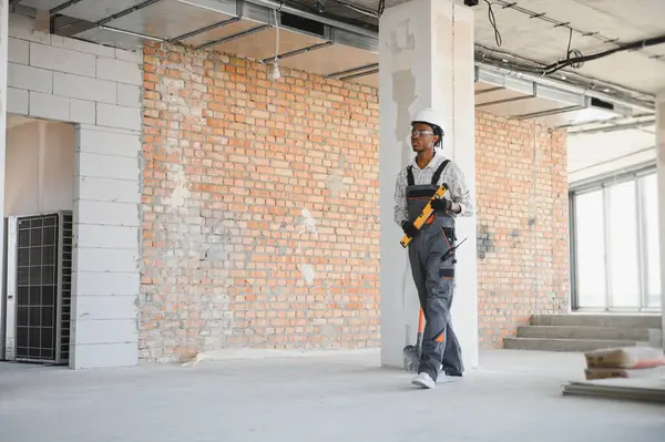 Afrika kökenli Amerikalı inşaat işçisi inşaat halinde, ruh seviyesini koruyor, iş tulumu, şapka ve eldiven giyiyor.