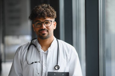 Genç Arap doktor modern bir hastane ortamında duruyor, profesyonellik ve uzmanlık yayıyor.