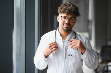 Beyaz önlük ve steteskop giyen genç Arap erkek doktor hastane koridorunda gülümsüyor, sağlık, çeşitlilik ve profesyonelliği temsil ediyor.