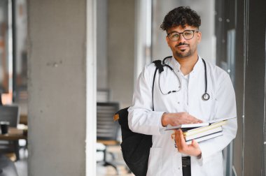 Kendine güvenen bir tıp öğrencisinin portresi kitap taşıyor ve üniversite koridorunda steteskop ve laboratuvar önlüğü giyiyor.