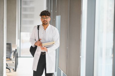 Kendine güvenen tıp öğrencisi kitap taşıyor, parlak bir üniversite koridorunda yürüyor, derslere hazır.