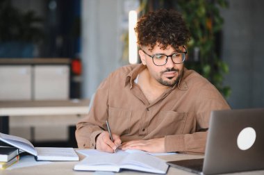 Genç Arap öğrenci evde veya kütüphanede dizüstü bilgisayar kullanarak çevrimiçi öğrenirken not defterine not alıyor