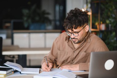 Üniversite kütüphanesinde okurken not defterine notlar yazan genç bir adam, yakınlardaki bir dizüstü bilgisayar ve ders kitaplarıyla öğrenmeye odaklanmış.