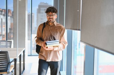 Genç Arap öğrenci kitap taşıyor ve modern bir üniversite binasında güvenle yürüyor, öğrenme ve büyüme olanaklarını kucaklıyor