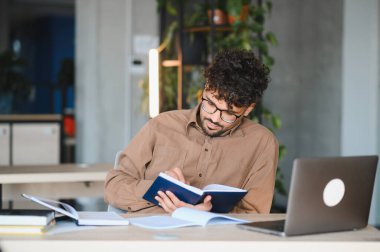 Genç bir Arap öğrenci, modern bir üniversite kütüphanesinde yaklaşan bir sınava çalışırken ders kitabından notlar alıyor.