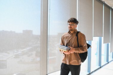 Üniversite koridorunda yürüyen genç bir Arap öğrenci, kitap ve sırt çantası taşıyor, özgüven ve kararlılık yayıyor.