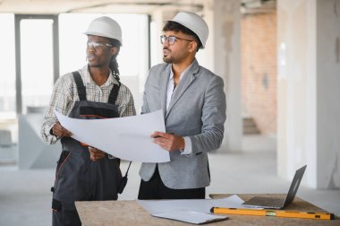 Güvenlik kaskı takan mimarlar ve inşaatçılar, hareketli bir inşaat sahasının planlarını yakından inceliyor ve proje detaylarını tartışıyorlar.