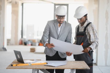 Şapkalı mimar ve müteahhit inşaat alanının planlarını inceliyor, bina planlarını tartışıyorlar.