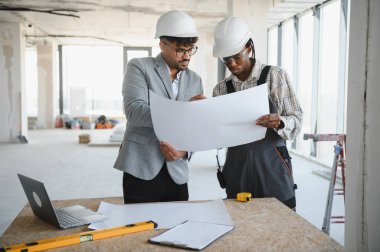 Her ikisi de başlık takan mimar ve müteahhit, inşaat sahasının geliştirme projesinde işbirliği yaparken ayrıntılı planları inceliyor.