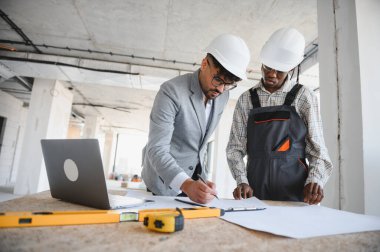 İnşaat alanının planlarını inceleyen mimar ve müteahhit, dizüstü bilgisayar ve ölçüm aletleri kullanıyor.