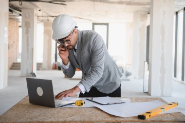 Arap mimar takım elbise ve güvenlik kaskı takıyor dizüstü bilgisayar kullanıyor ve inşaat alanında telefonla konuşuyor.