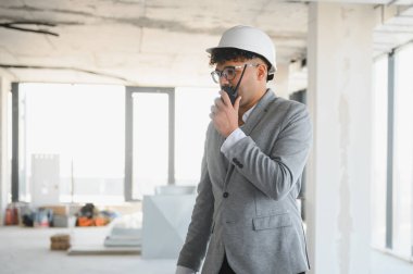 Arap mimar takım elbise ve şapka giyiyor. Telsiz kullanıyor. İnşaat halindeki bir binanın içindeki operasyonları koordine ediyor.