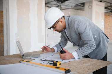 Arap mimar inşaat sahasında çalışırken, dizüstü bilgisayar ve telsiz kullanarak, planları kontrol ederek ve notlar alarak,
