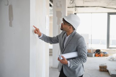 Arap mimar inşaat alanının planlarını elinde tutarken ve inşaat alanını incelerken duvara işaret ederken,