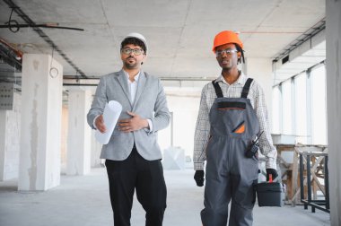 Elinde planlar olan mimar inşaat halindeki bir binanın içinden alet çantası taşıyan bir inşaatçıyla yürüyor.