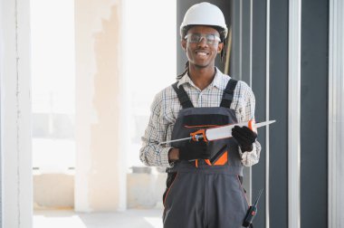 Gülümseyen Afro-Amerikan inşaat işçisi elinde silikon dolgu tabancasıyla güvenlik kaskı takıyor ve iş tulumu giyiyor.