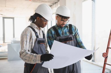 Güvenlik kaskı ve eldiven takan iki müteahhit bir inşaat alanının planını inceliyor.