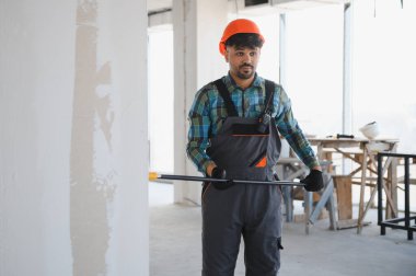 Arap mühendis turuncu bir kask takıyor ve inşaat alanında çalışırken elinde bir ölçüm aleti tutuyor.