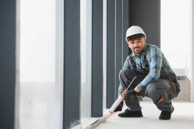 İnşaat işçisi, yoğun bir inşaat sahasındaki pencere çerçevesine dolgu macunu sürüyor, her detayı kalite ve hassasiyet sağlıyor.