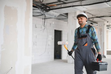 Arap müteahhit tulum giyiyor ve kafasında boya silindiri ve inşaat alanında yürüyen alet çantası taşıyor.