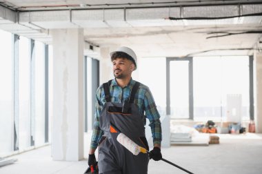 Arap mühendis, inşaat halindeki binanın içinde yürürken şapka takıyor. Boya silindiri ve alet çantası taşıyor.