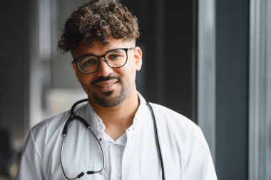 Kendine güvenen, gülümseyen, gözlük takan, steteskop takan, hastane ortamında profesyonelliği ve uzmanlığı temsil eden Arap erkek doktor.