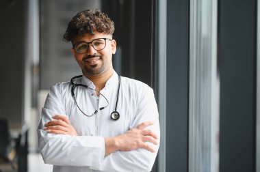 Kollarını kavuşturup gülümseyen, modern hastanede beyaz önlük ve steteskop giyen kendine güvenen Arap erkek doktor.