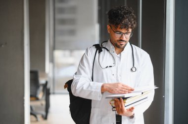 Genç Arap tıp öğrencisi gözlüklü ve beyaz önlüklü üniversitede kitap ve sırt çantası taşıyor.