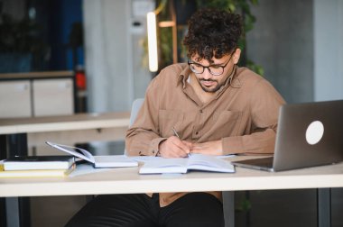 Genç Arap öğrenci, kitap ve kaynaklarla çevrili bir üniversite kütüphanesinde özenle çalışırken bir deftere notlar yazıyor.