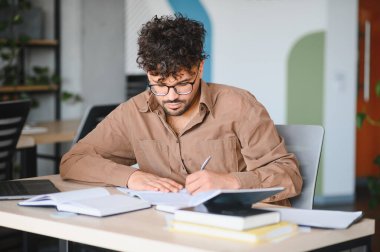Genç Arap öğrenci, kitaplarla ve doğal ışıkla çevrili bir üniversite kütüphanesinde notlar alıyor ve özenle ders çalışıyor.