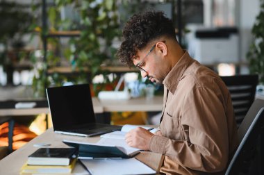 Genç Arap öğrenci dizüstü bilgisayar ve kitaplarla üniversite kütüphanesinde okurken deftere notlar yazıyor.