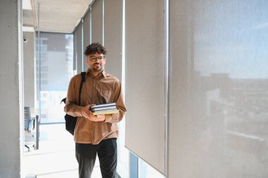 Genç Arap öğrenci üniversite koridorunda yürüyor, elinde kitaplar ve sırt çantasıyla, akademik uğraşlara dalmış ve öğreniyor.
