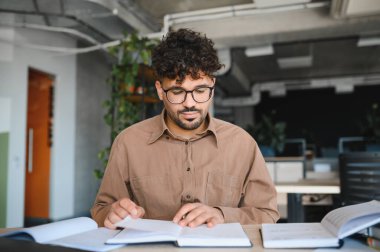 Kütüphanede okuyan ve okuyan, sınavlara hazırlanan ve akademik projeleri için araştırma yapan konsantre genç bir adam.