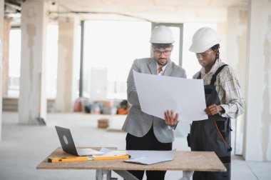 Bir inşaat alanının planlarını inceleyen mimar ve inşaatçı, proje detayları ve işbirliği hakkında tartışmalara giriyor