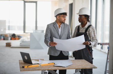 Mimar ve müteahhit başlık takıyor, planları tartışıyor ve inşaat halindeki bir binanın planları üzerinde işbirliği yapıyor.