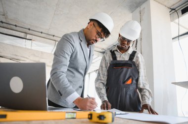 İnşaat alanında birlikte çalışan mimar ve müteahhit, planları gözden geçirir ve dizüstü bilgisayar kullanır.