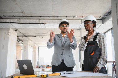 Güvenlik kaskı takan mimar ve müteahhit tamamlanmamış binada el hareketleriyle inşaat projesini tartışıyor.