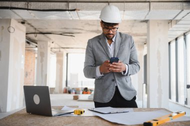 İnşaat alanında akıllı telefon kullanan takım elbiseli ve kasklı bir Arap mimar. Üzerinde planlar, dizüstü bilgisayarlar ve ruh seviyesi olan bir masa var.
