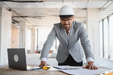 Kasketli genç bir mimar, planları gözden geçirir ve hareketli bir inşaat sahasında dizüstü bilgisayar üzerinde çalışır, güven ve profesyonellik yayar.