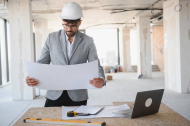 Arap mimar, inşaat alanında dizüstü bilgisayar ve aletlerle masaya otururken başlık takıyor ve planı inceliyor.