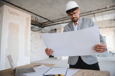 İnşaat alanının planını inceleyen Arap mimar, şapka ve takım elbise giyiyor, dizüstü bilgisayarı ve panosu var.