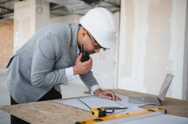 Arap mimar takım elbise ve şapka takıyor, telsiz kullanıyor ve inşaat alanının planlarını inceliyor.