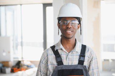 Afrika kökenli Amerikalı inşaat işçisi baret ve güvenlik gözlüğü takarak parlak bir şantiyede gülümsüyor.